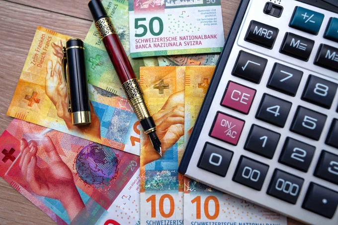Auf dem Bild sieht man verschiedene Schweizer Banknoten, einen Taschenrechner und zwei Kugelschreiber auf einem Tisch