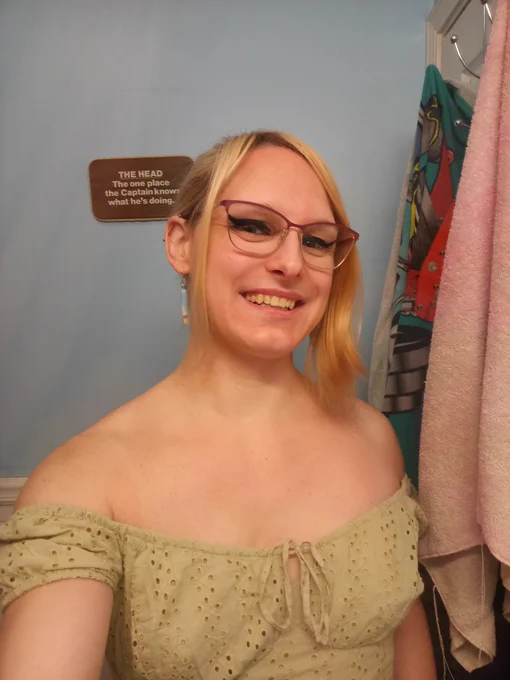 A woman in a light and airy green puffy sleeve top with her wide and warm smile framed by long blonde curtain bangs. She has a blue popsicle earring.