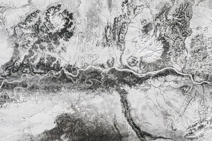 A frozen river winds from east to west across the image, appearing bright white. Nearby meandering channels are also frozen, and much of the surrounding land is snow-covered, but areas of light brown peek through the snow, especially surrounding the river. 
