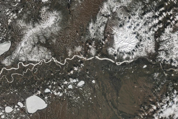 A river winds from east to west across the image, brown-white ice stretches most of the river space, with breaks of open space where ice is broken up. Most of the surrounding land is snow-free, appearing dark green and brown. 
