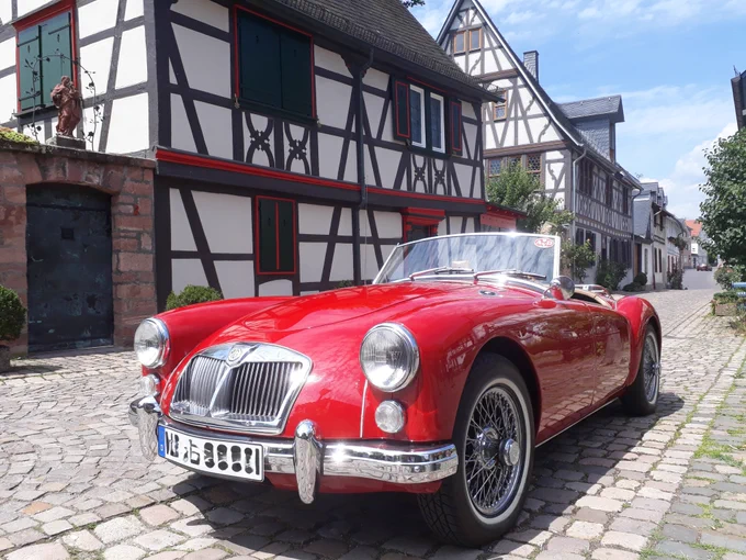 Zu sehen ist ein knallrotes Oldtimer-Cabriolet. Man sieht die Vorderfront des Autos mit großen runden Scheinwerfern und einem silbernen Kühlergrill. Das Auto wurde von schräg vorne fotografiert. Es steht in einer Kopfsteinpflastergasse. Im Hintergrund sind Fachwerkhäuser zu sehen. Es ist sonnig.