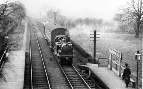 Looking down onto Bandon Halt, the name board legible on the up platform as a train approaches and a solitary passenger on the platform. In the photograph the platforms look fairly new and clean suggesting the photograph was taken soon after opening. The photographer was facing west, i.e. the train is travelling from Wallington to Waddon with the photographer standing on the Plough Lane bridge. There is a motor train with an LBSCR ballon coach in tow possibly powered by TerrierNº661