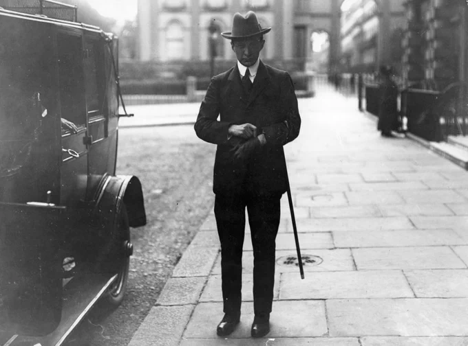 A person in formal attire stands next to a vintage car holding a cane, with buildings and a fence in the background.