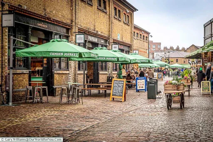 The cobbled courtyard of the North Yard of Camden Market, home to artisan food stores with parasol covered benches outside.