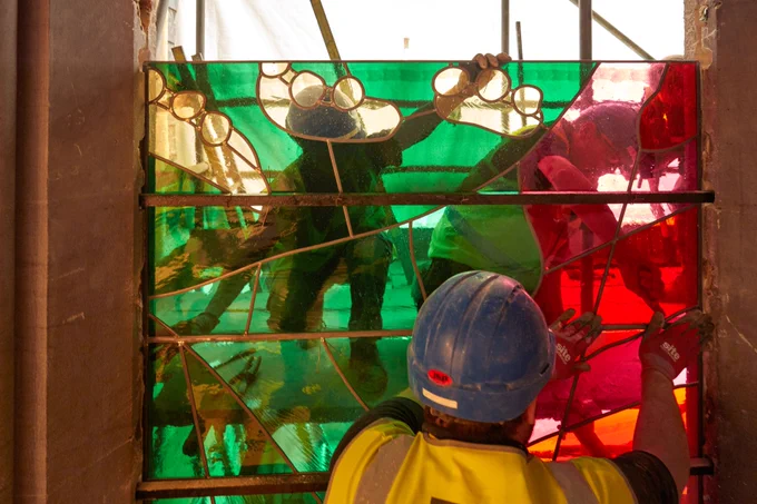 A colourful panel of stained glass being installed in Westminster Abbey by workers wearing hard hats and high viz jackets