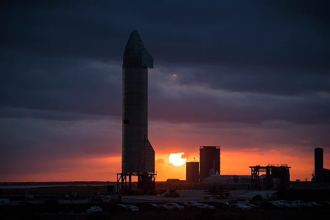 A large rocket stands on a launch pad at sunset, silhouetted against an orange sky with scattered clouds and parked vehicles nearby.
