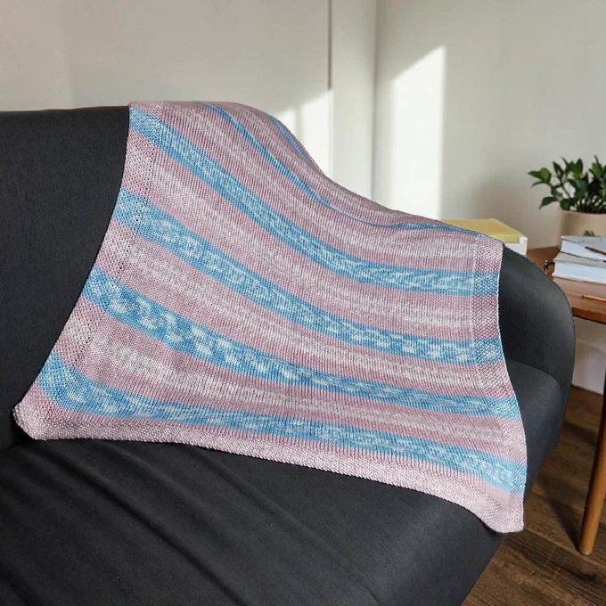 A hand knitted blanket with  stripes in pastel pink, light blue, and white drapes over the arm/back of a dark grey fabric sofa. Background: plain light off-white walls, bright sunlight streaming in, wooden floor, side table with books/notebook, pen, and a potted green plant in a beige pot. Calm, bright room setting.