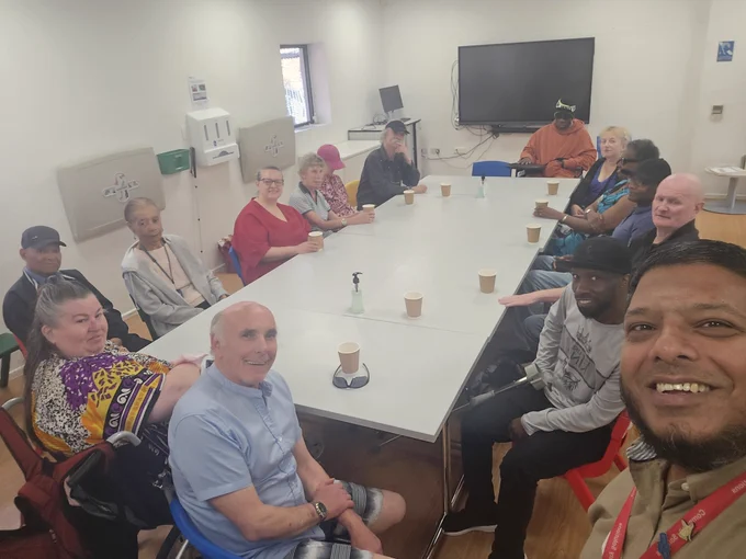 A group of people is seated around a large rectangular table in a well-lit room with white walls and a wooden floor. There are approximately fourteen individuals, each sitting on colorful chairs, some smiling at the camera. Among the attendees, there are a mix of men and women, varying in age. On the table, there are several disposable coffee cups, and a hand sanitizer bottle is also visible. A large television screen is mounted on the wall in the background, and there are some wall-mounted storage units and a window on the left side of the room.