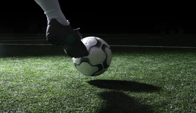 A football boot about to make contact with a white and black football on an illuminated green artificial grass pitch at night, shot from a low angle with dramatic lighting against a dark background.