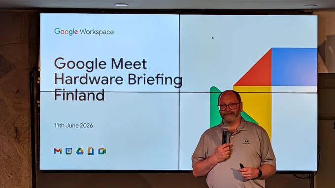 A photo of a man standing in front of large screens, holding a microphone as he presents. On the screens are the words “Google Meet Hardware Briefing Finland” and the date of the event.