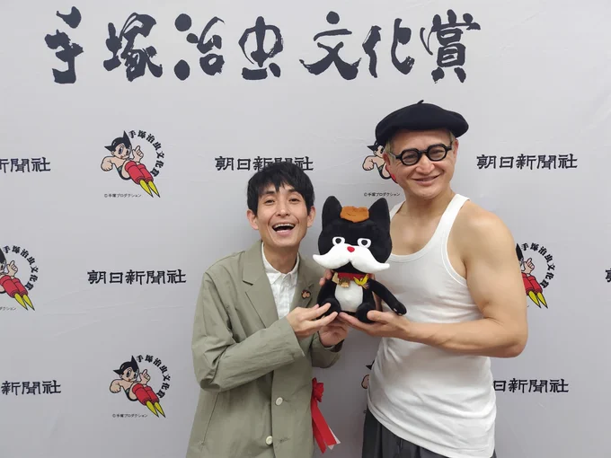 矢部太郎さんと毛塚治虫先生が、朝日新聞社オリジナルキャラクターのマダニャイを手に、記念撮影をしている写真です。