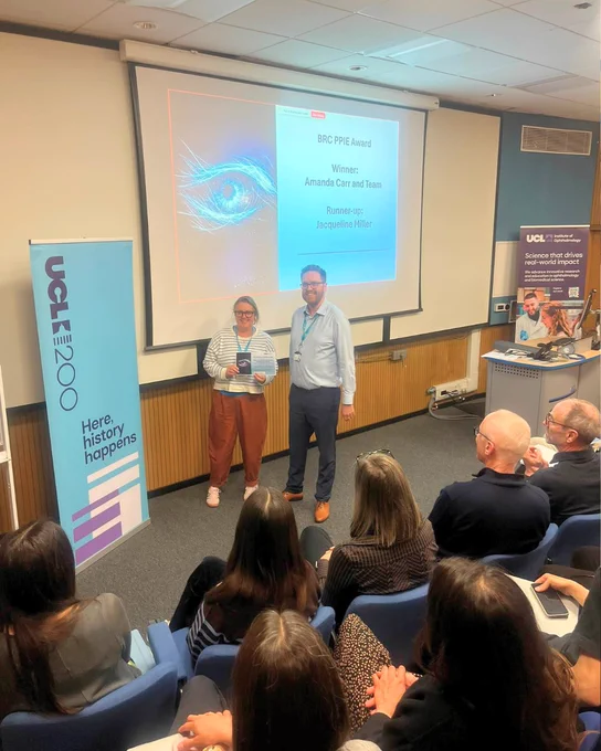 Person holding a certificate stands beside another person at the front of a lecture room during an awards presentation. A projected slide behind them reads “BRC PPIE Award” with the winner and runner-up names. An audience seated in rows faces the presenters. A vertical banner on the left reads “ECRF 2020 – Here, history happens,” and a UCL Institute of Ophthalmology banner is visible on the right.