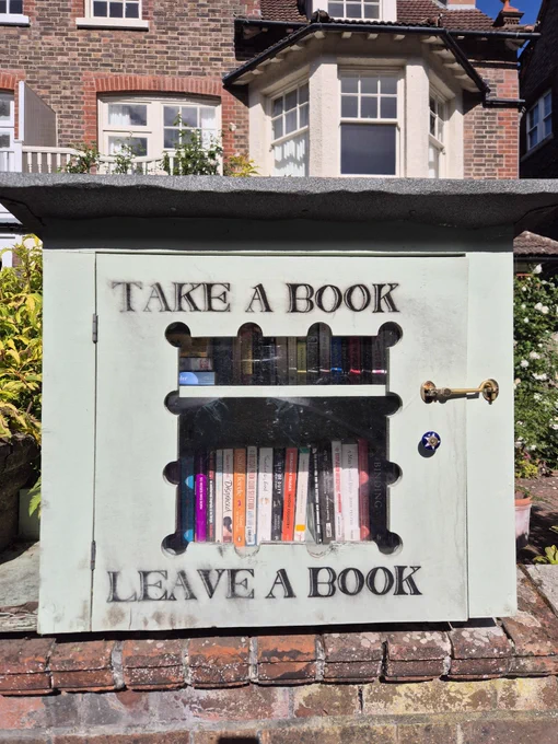 Book cabinet fixed to a garden wall, filled with books. The words, Take a Book, Leave a Book, stencilled on the front.