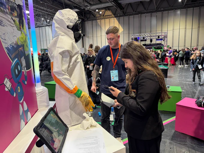 A person in protective gear stands at an exhibition booth with colorful decorations, including a cartoon robot. Another attendee holds a device and engages with the display. The event is busy with many people in the background.