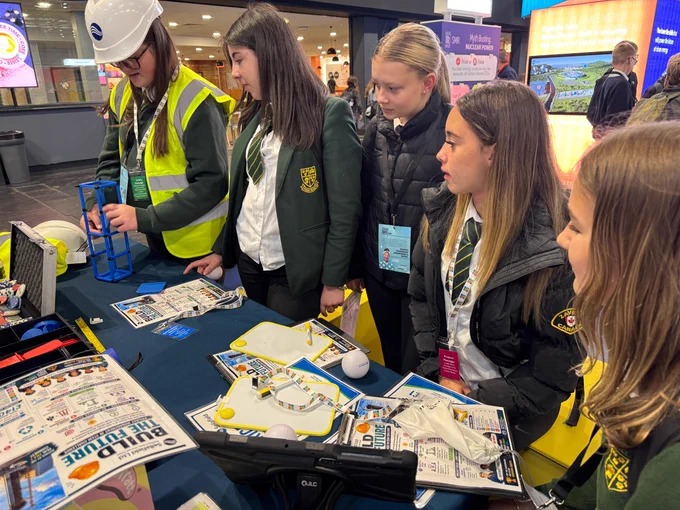 A group of people in school uniforms attentively observe a person in a high-visibility vest and hard hat demonstrating a small structure. The display table is covered with pamphlets titled "Build the Future" and educational materials.