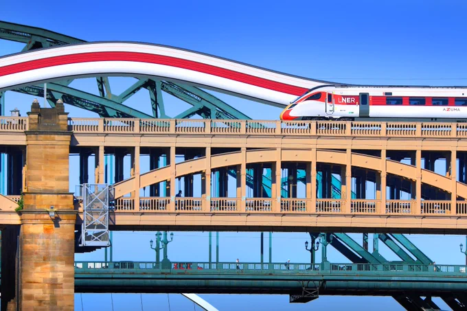 The end of an Azuma train on Newcastle's High Level Bridge, with a slice of the train's livery colours extending in line with its red stripe and arching over like another bridge. The Tyne Bridge is in the background.
