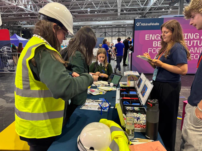 People are gathered around a table at an event. One person is wearing a hard hat and reflective vest. The table is filled with various stationery items.