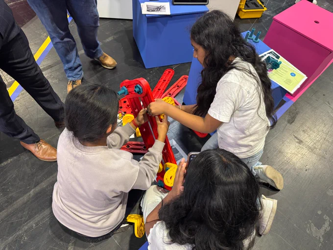 Three people are sitting on the floor, engaging with a colorful construction set, building with large interlocking pieces in red, yellow, and blue. Other individuals stand nearby on a dark floor.