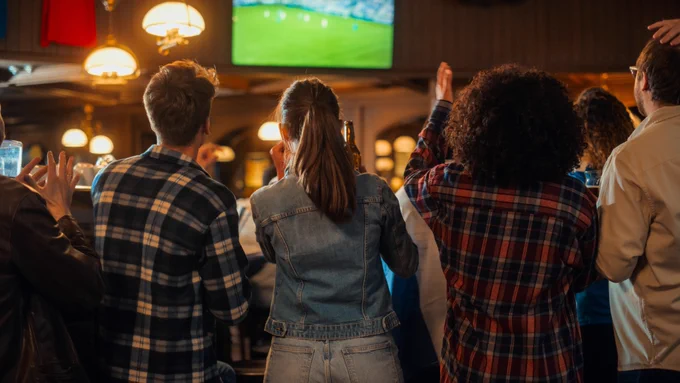 People in a pub watching a football match on a screen, with some raising their hands.