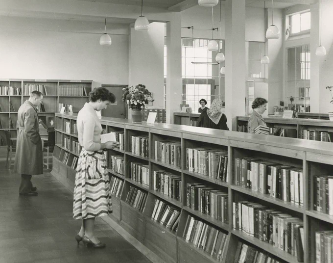 A library interior. Several long rows of shelves in the foreground of the photograph and another running along the backwall in the mid background of the photograph. Two figures inspecting the row of bookshelves in the immediate foreground. The female figure in the forefront is holding a book. The two female figures perusing the second row of shelving have their backs to the camera. The figure in the foreground is holding a large pile of books. The figure in the background is wearing a headscarf. The figure in the far top right background is looking on towards the shelving and customers.  