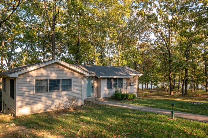 A one-story house with beige siding is surrounded by tall trees with green leaves. A paved path leads up to the front door, and a grassy lawn extends in front. Located in a peaceful, wooded area near a body of water visible in the background.