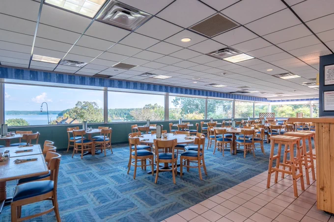Spacious dining area with large windows offering a scenic view of trees and a lake outside. The room is filled with wooden tables and chairs, arranged neatly on a blue carpet. Natural light brightens the space.