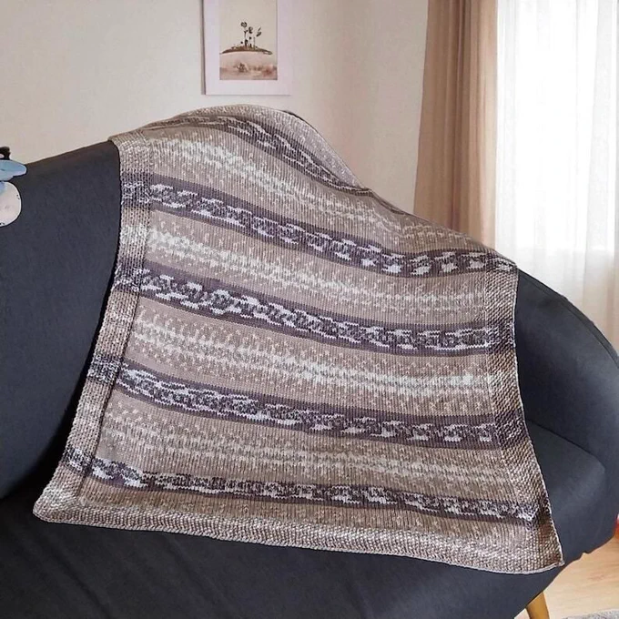 A knitted blanket in shades of brown, beige, and white, draped over a dark grey sofa. The blanket features horizontal stripes with textured patterns. The neutral colour palette and soft texture suggest a cosy, home decor item.