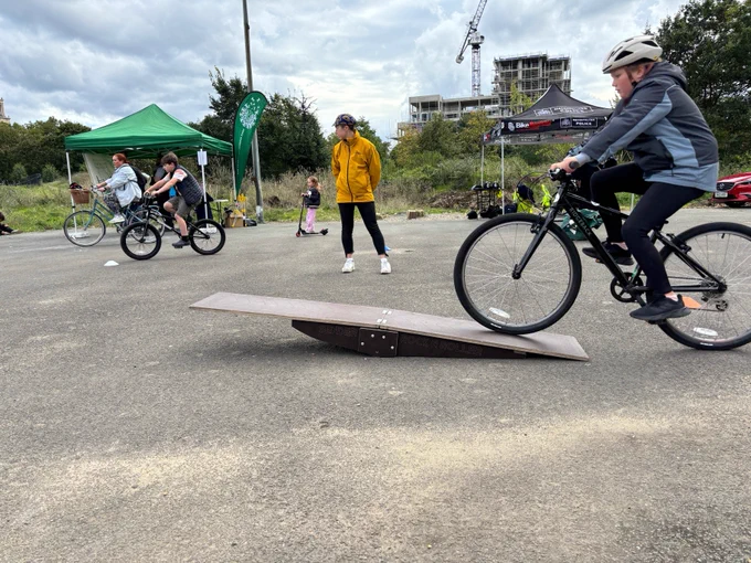 Children and adults at an outdoor cycling event, with one child riding a bike up a ramp.