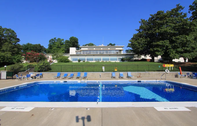 A large rectangular swimming pool with clear blue water is in the foreground, surrounded by a paved area with lounge chairs. In the background, there is a mid-century modern style building, situated on a grassy hill, with large trees and a bright blue sky above.