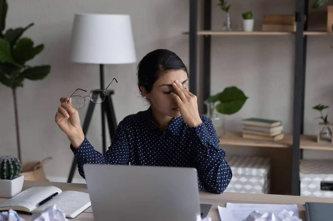 A tired woman sitting at her desk in front of a laptop, holding her glasses and pinching her nose, illustrating workplace stress, meeting fatigue, and burnout.