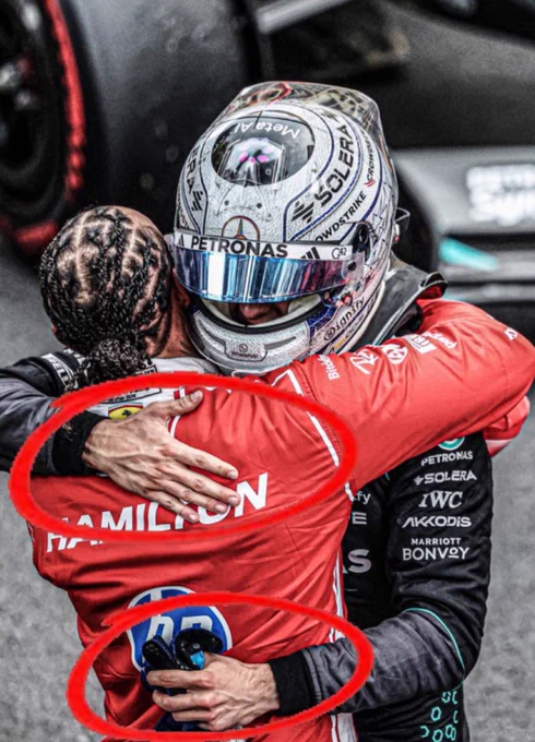 George Russell in Mercedes AMG Petronas F1 race suit and helmet embraces Lewis Hamilton, who wears a red Ferrari race suit with HAMILTON printed across the back, in parc ferme after a Formula 1 Grand Prix. Red circles highlight Russell’s hands on Hamilton’s back, one still holding his racing gloves. Mercedes sponsors Petronas, IWC, Solera and Akkodis visible on the suit.