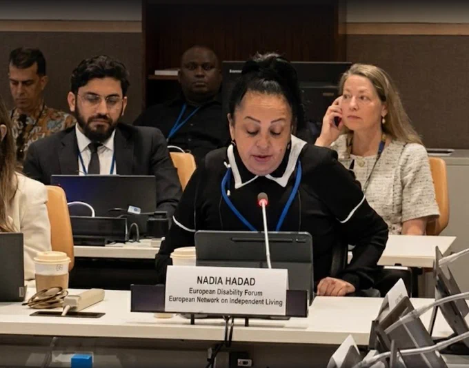 Alt text: Nadia Hadad speaks into a microphone at a COSP19 side event, seated behind a nameplate reading “Nadia Hadad, European Disability Forum, European Network on Independent Living.” Other participants are seated behind her in the meeting room.