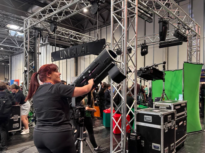 An individual adjusts a large stage light within an indoor exhibition space. Surrounding the area are metal trusses, stacked equipment cases, and a green screen setup. A sign reads "Unusual." Several people are visible in the background, engaging with the displays.