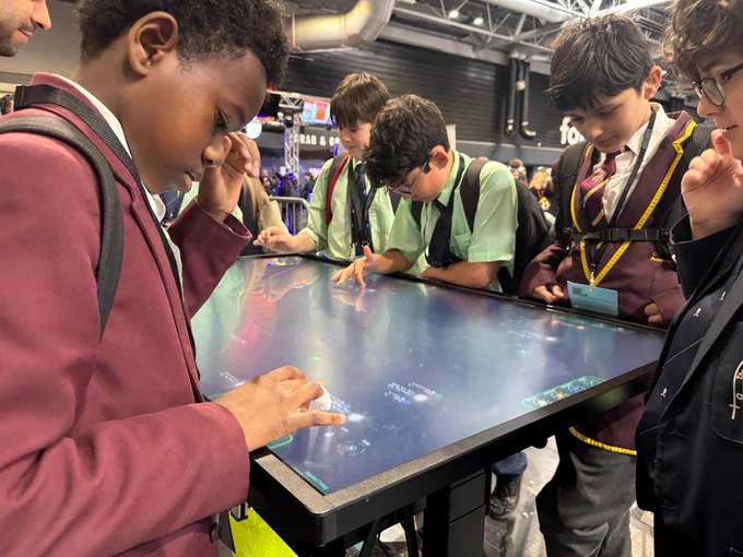 A group of people are gathered around a large interactive touchscreen table at an event. They are engaged with the display, using their hands to interact. The background shows part of a crowded venue with various booths and people.