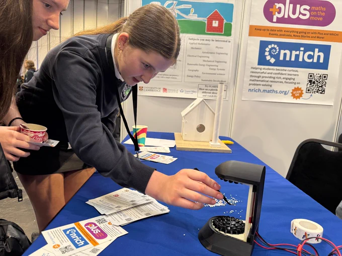 Two people are engaged in an interactive exhibit at a mathematics event. One person is dropping small white objects into a machine. Promotional materials for "Plus Maths" and "NRICH" are visible on the table, along with informational brochures. A QR code can be seen on a poster in the background.