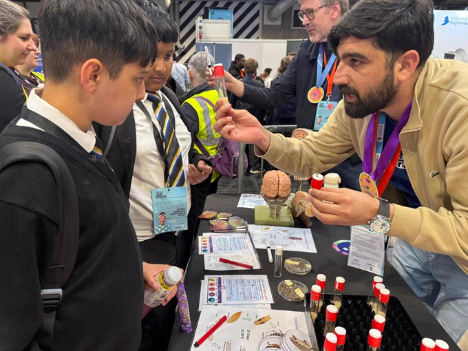 A person at a science fair booth enthusiastically explains an exhibit to two attentive attendees. The table features anatomical brain models and various educational materials. Visitors and a busy exhibition environment are visible in the background.