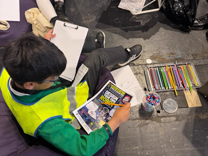 A person wearing a yellow vest sits on a purple bean bag, holding a flyer titled "Design a Robot" and a clipboard with paper. Nearby on the floor are coloured pencils and markers.