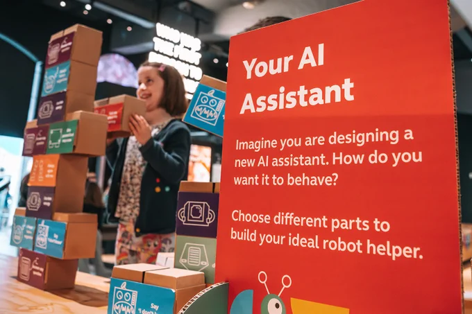 Child taking part in the 'Demystifying AI' programme at We The Curious science centre in Bristol. Text on signage says 'Your AI Assistant' Imagine you are designing a new AI assistant. How do you want it to behave? Choose different parts to build your ideal robot helper.