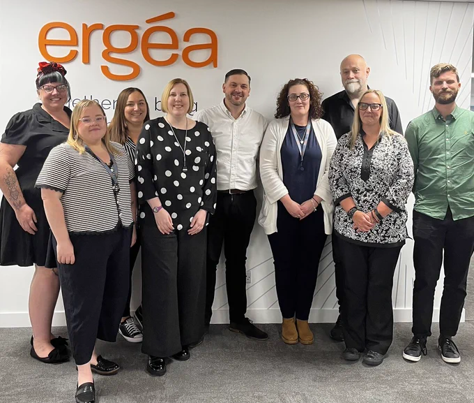 Group of nine professionals standing in front of a white wall with the orange 'ergéa' logo, dressed in business casual attire.