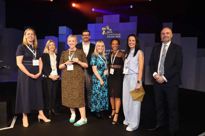 A group of people are standing together on a stage at the NHS Excellence Awards 2026. They are dressed in formal attire and appear to be posing for a photo, some holding an award. The background features the event's logo.