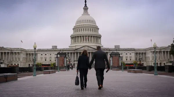 A still from the film 4000 Days. Seen from the back, a man and woman hold hands as they walk towards the US Capitol building, a large white public edifice topped with a dome.