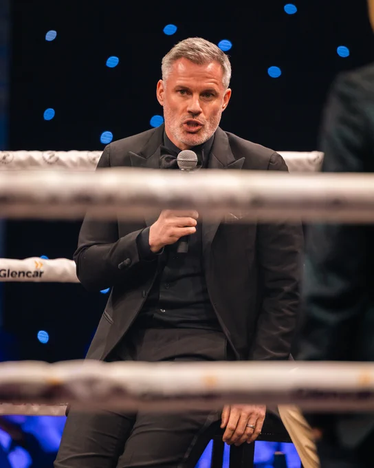 A well-dressed man in a tuxedo is speaking into a microphone while sitting inside a boxing ring. The scene appears professional and formal.