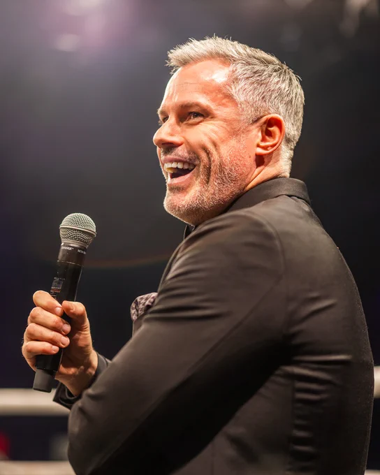 A man with gray hair and a black suit holds a microphone while speaking energetically. The lighting is warm and highlights his expressive demeanor.
