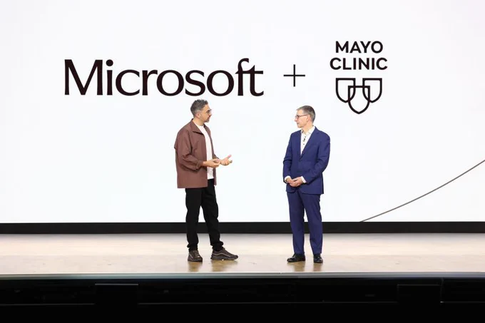 Two speakers stand on a stage in front of a large screen displaying the Microsoft and Mayo Clinic logos, facing each other as they talk during a presentation.