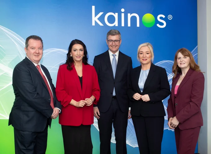 Image of five people standing in front of Kainos branding. Included are First Minister Michelle O’Neill, deputy First Minister Emma Little-Pengelly, Economy Minister Dr Caoimhe Archibald, Brendan Mooney (CEO, Kainos) and Kieran Donaghue (CEO, Invest NI).