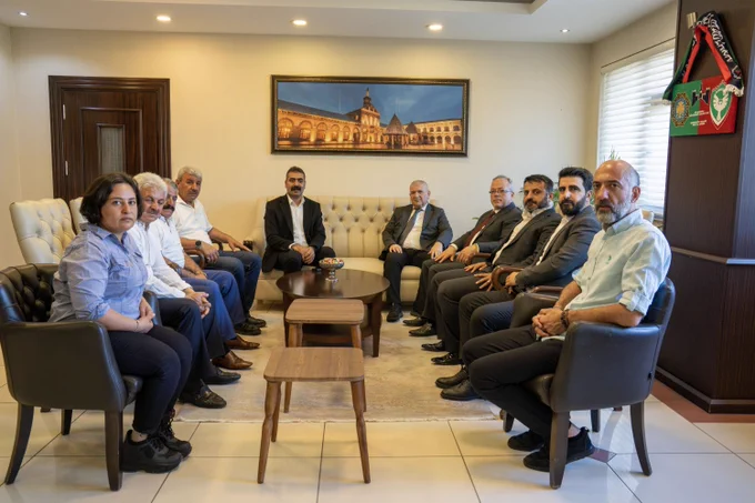 Bir ofis ortamında, iki sıra halinde karşılıklı oturan 11 kişi ve arka bölümde bir koltukta oturan bir kişi görülüyor. Ortada sehpa ve küçük masalar yer alıyor. Duvarlarda çerçeveli bir yapı fotoğrafı bulunuyor. Sağda pencere ve dolap, solda kapı görülüyor. Katılımcılar kameraya dönük şekilde oturuyor.