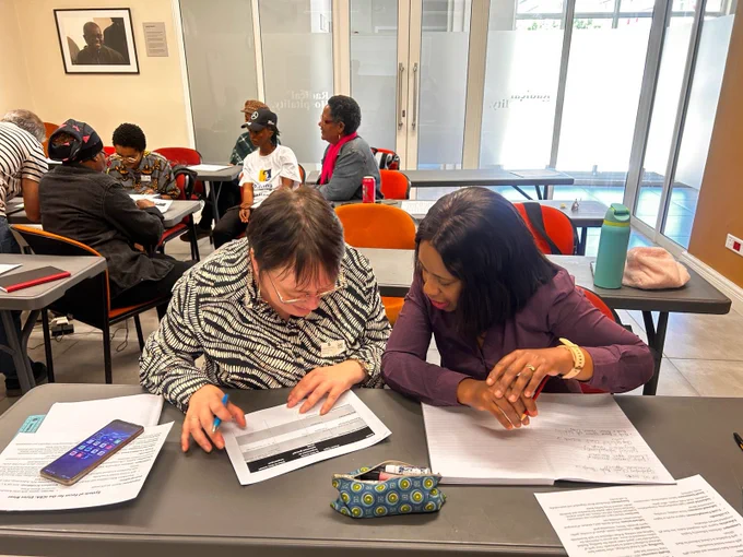 A group of people sit in a classroom or training space, working together at tables. In the foreground, two individuals focus on printed materials and notes, writing and discussing their work. Other small groups are visible in the background, also engaged in discussion and activities. The room has desks, chairs, notebooks, and personal items such as a phone and water bottle on the tables. A framed photograph hangs on the wall, and glass doors are visible at the back of the room, giving the space a professional, workshop-like setting.  