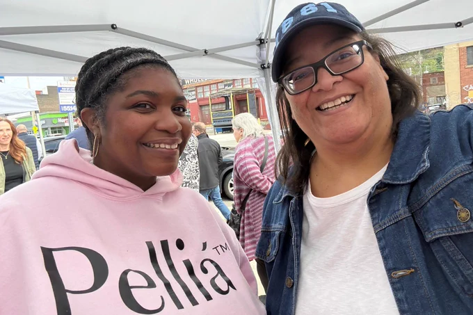 Two women smiling and posing for a selfie outdoors under a canopy, with other people and buildings visible in the background.
