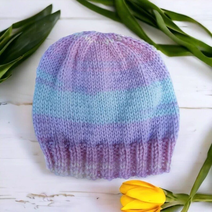 A hand knitted baby hat in pastel shades of pink, lilac, green and yellow on a wooden surface with a yellow flower
