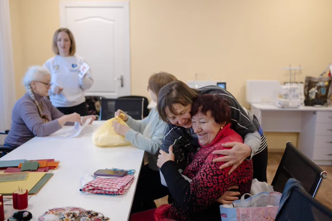 Participants of the Leisure and Social Services Centre for Older People in Synelnykove take part in a crafts session. Two women share a warm embrace while other participants work on handmade projects, reflecting the centre’s role in fostering social connections among local residents and internally displaced people.
